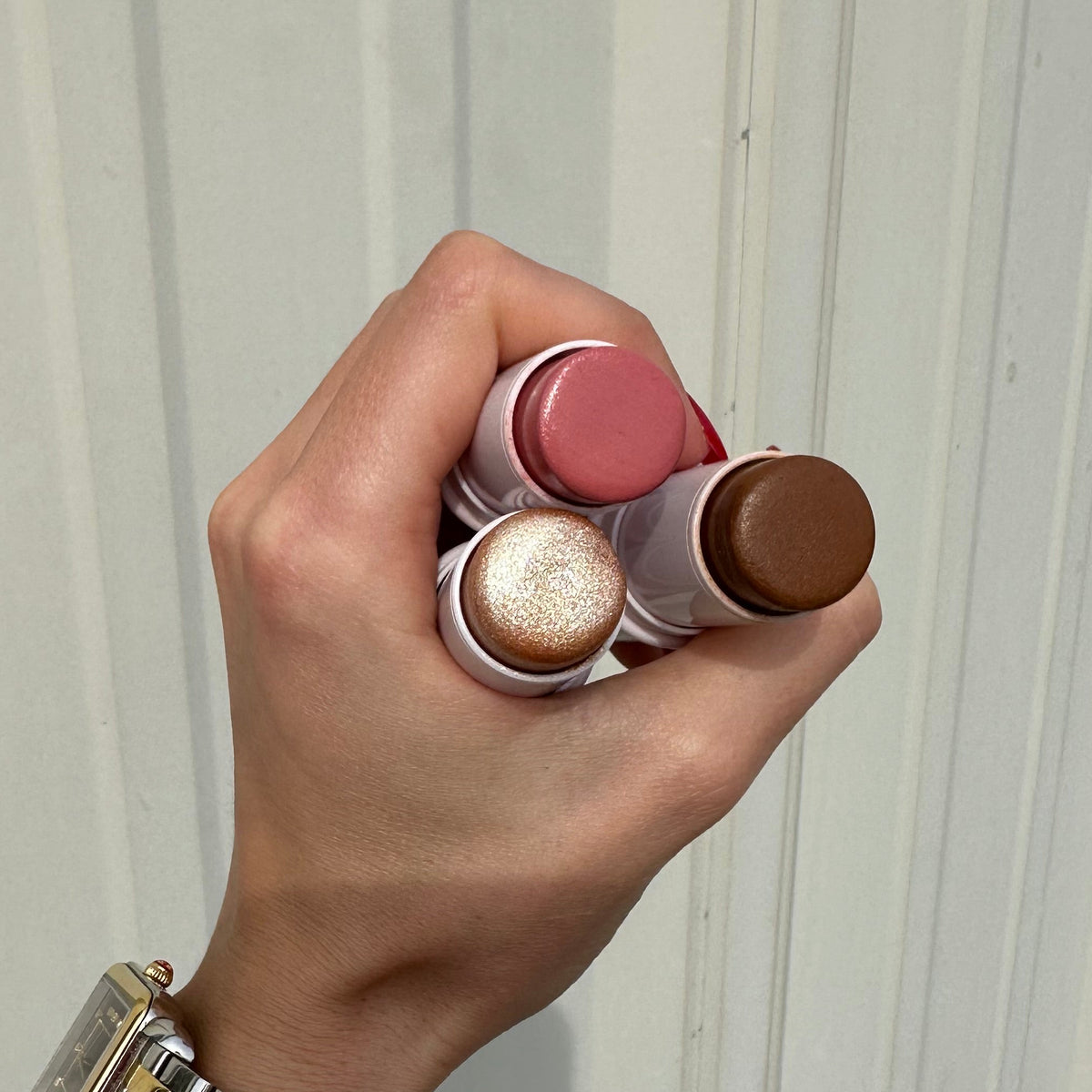 A hand holding three cosmetic stick products, Hope Beauty Radiant Sticks, including a pink stick (I Am Honored), a shimmery champagne stick (I Am Radiant), and a brown stick (I Am Resilient), against a white paneled background. Makeup inspired by affirmations. Inspirational Makeup. Christian Makeup.
